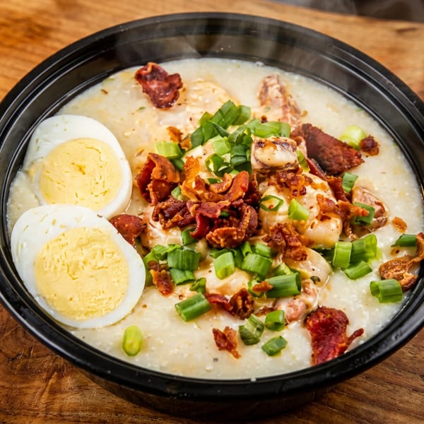 Steaming bowl of creamy grits topped with shrimp, bacon, green onions, and two hard-boiled egg halves.