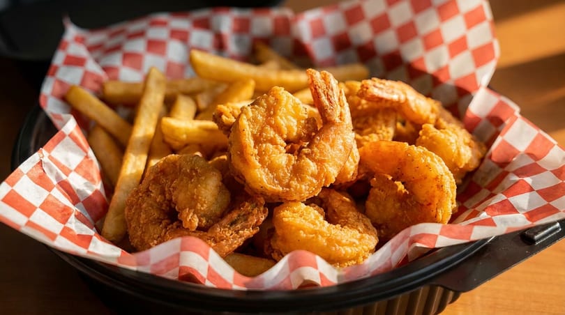 Golden fried shrimp and french fries in a black basket lined with red and white checkered paper.